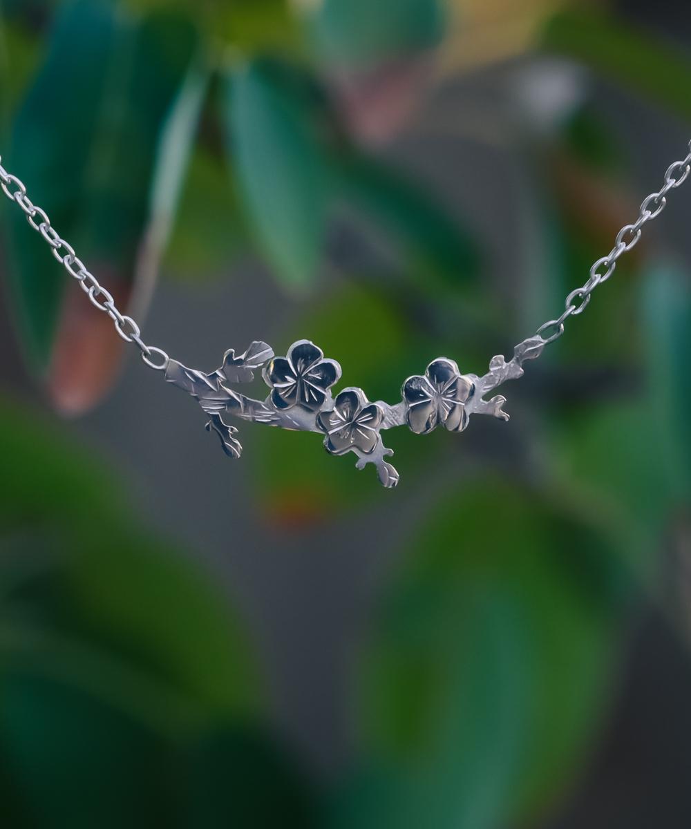 Silver necklace in the shape of a branch with three cherry blossoms. Button in top left of image reads FLORAE and will take you to plant themed items